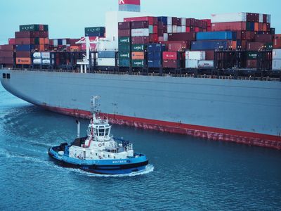 A photo of a small blue and white tugboat next to a very large shipping container ship full of multi-colored shipping containers.